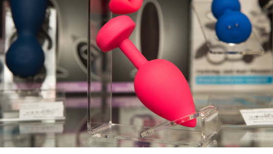 A range of silicone sex toys in glass cases being shown. Main colours are deep blue from a silicone dildo and hot pink from a silicone butt plug