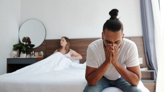 Man with ED (erectile dysfunction) sat on the edge of the bed with his girlfriend lying in the bed behind